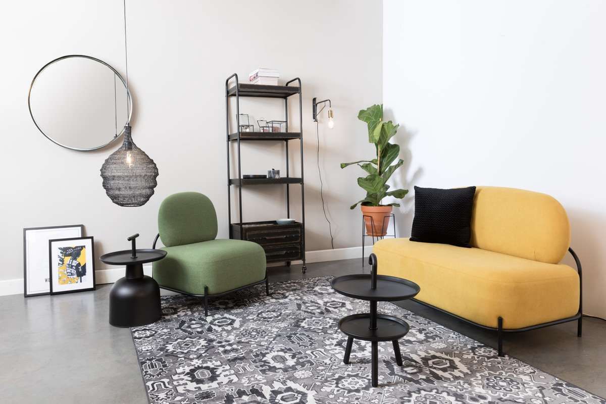 Grafika przedstawia salon, w którym znajduje się żółta sofa oraz fotel w kolorze zielonym. Na szarej podłodze umieszczono wzorzysty dywan, natomiast na ścianie jest lustro oraz regał.
