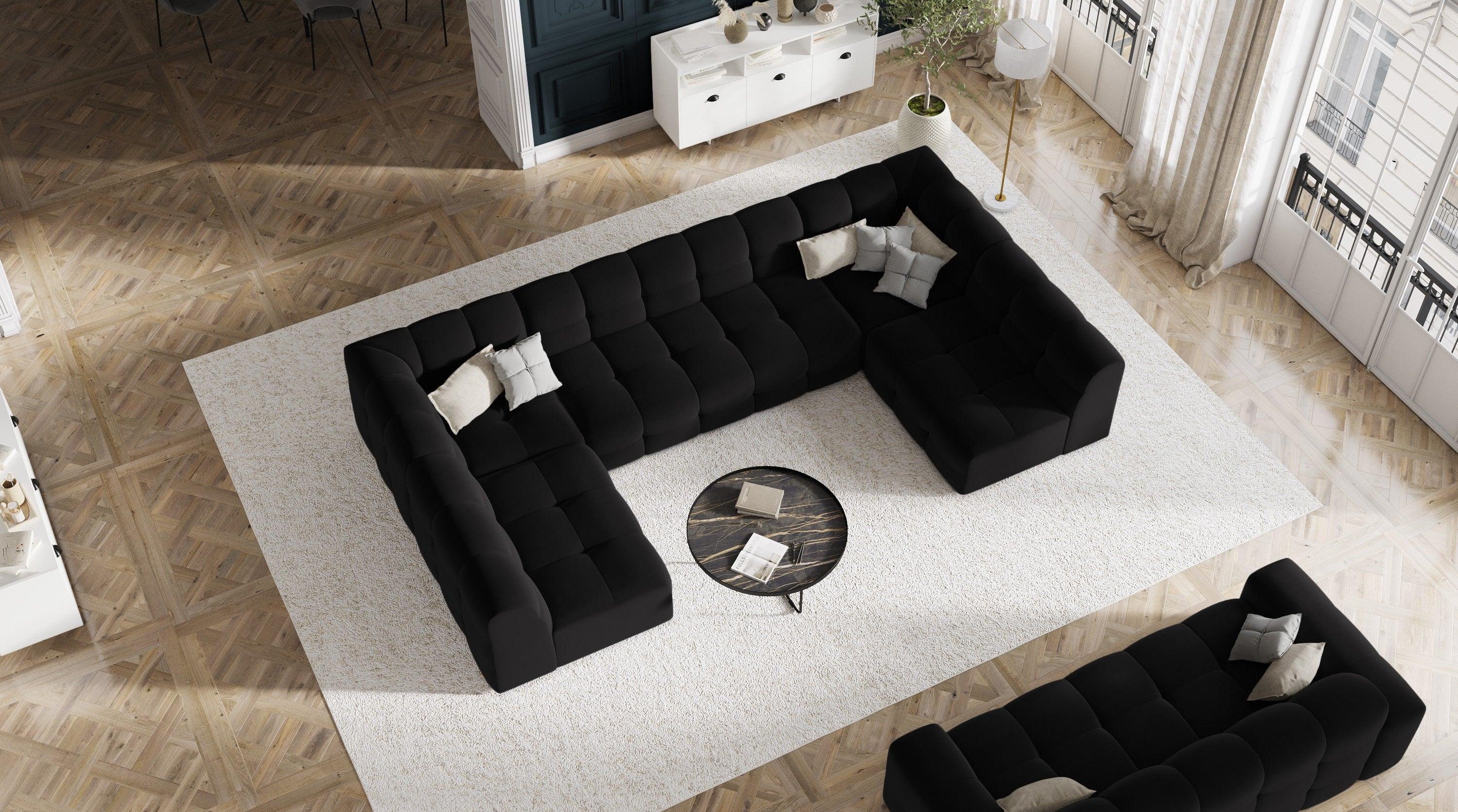 Elegancki narożnik w kształcie litery U w kolorze czarnym stoi na prostokątnym, beżowym dywanie wykonanym z miękkiej tkaniny. Naprzeciw narożnika znajduje się sofa z tej samej kolekcji. Pomiędzy nimi umieszczono okrągły stolik kawowy. 