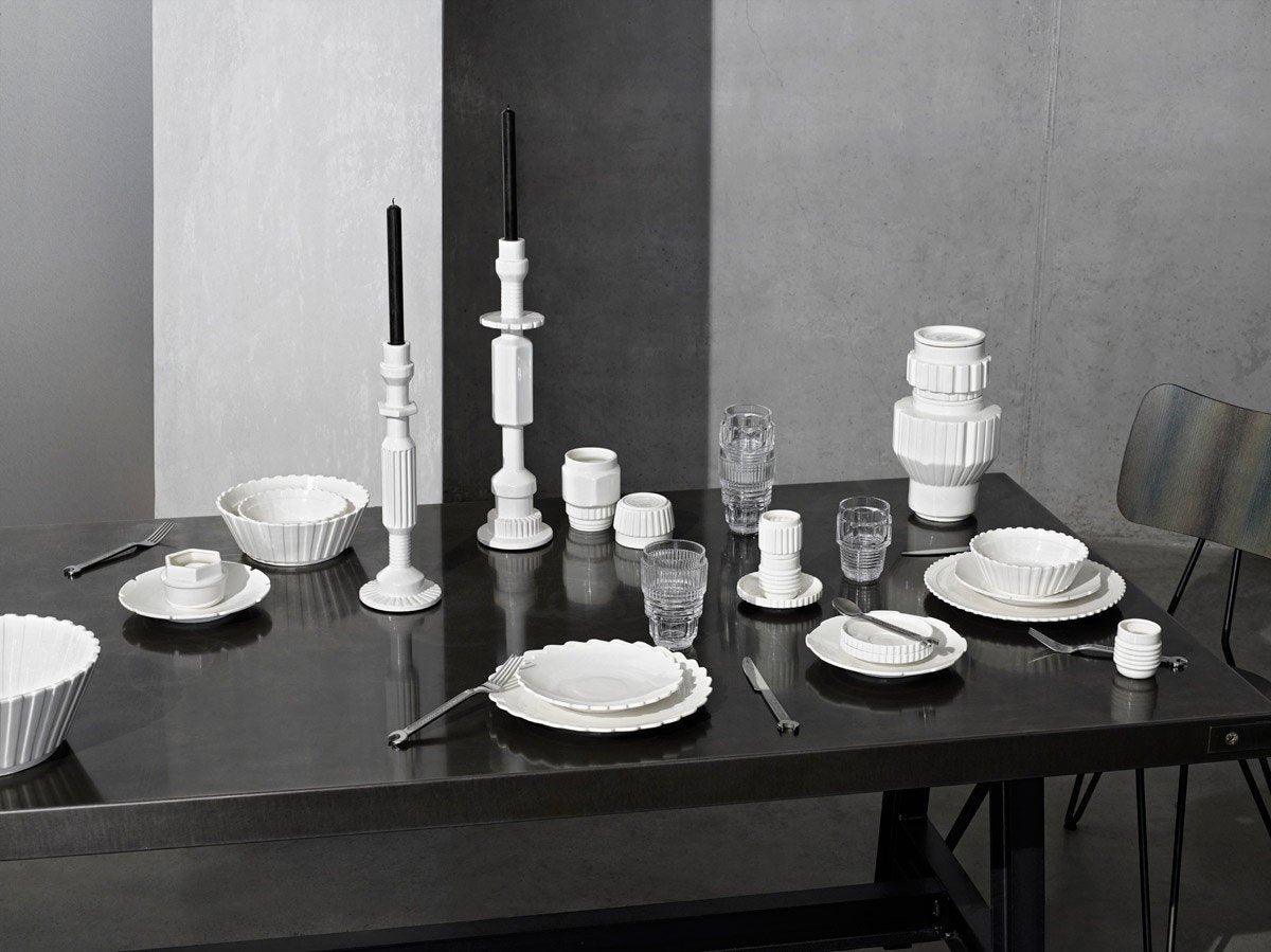 Czarno białe zdjęcie przedstawia elegancko zastawiony stół, na którym znajdują się talerze, sztućce oraz świeczniki i wazon. Monochromatyczność dodaje grafice klasy i elegancji.