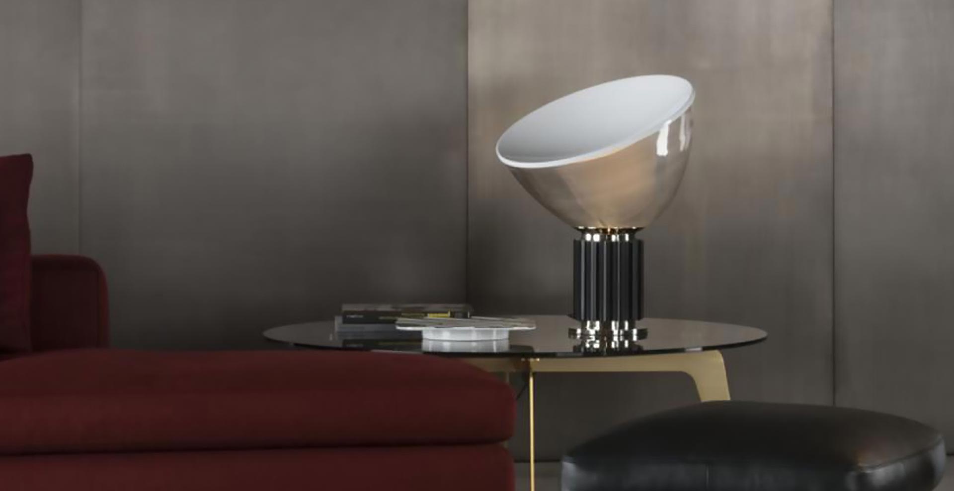 Minimalistyczna lampa stołowa, której klosz przypomina wygiętą misę stoi na stoliku bocznym obok bordowej sofy. Pomieszczenie jest urządzone w nowoczesnym, brutalnym stylu.